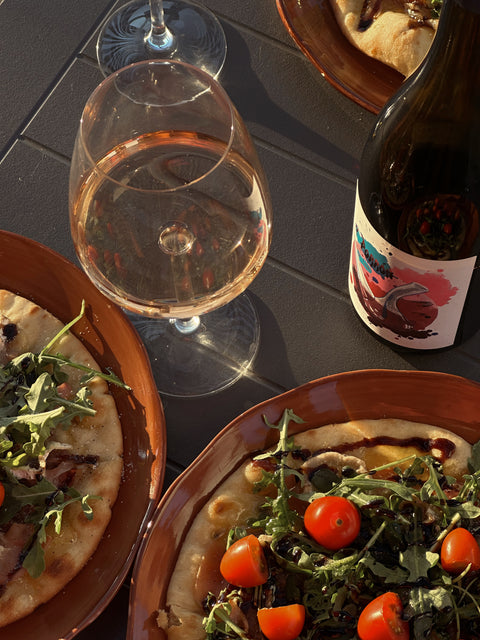 Two pizzas with tomatoes and greens on a table with a glass of wine and a bottle.