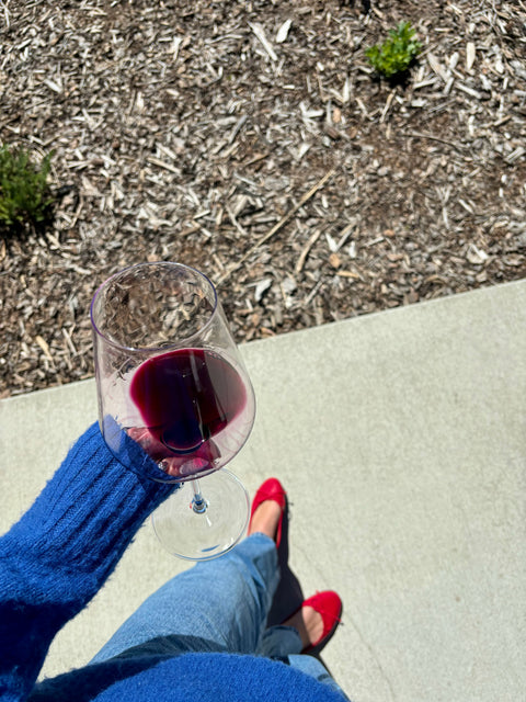 Person holding a glass of red wine on a concrete surface with mulch and plants in the background